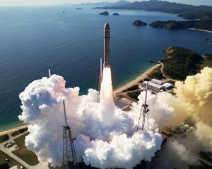 Rakieta nośna wznosi się pionowo z nadmorskiego kompleksu startowego, pozostawiając jasny słup ognia i gazów spalinowych; poniżej gęsta chmura białego dymu rozlewa się nad platformą startową, a w tle widoczne są ocean, linia brzegowa i tereny leśne.