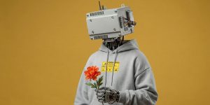 Humanoidalny robot z prostokątną mechaniczną głową w jasnoszarej bluzie z kapturem, trzymający pomarańczowy kwiat na żółtym tle studyjnym.